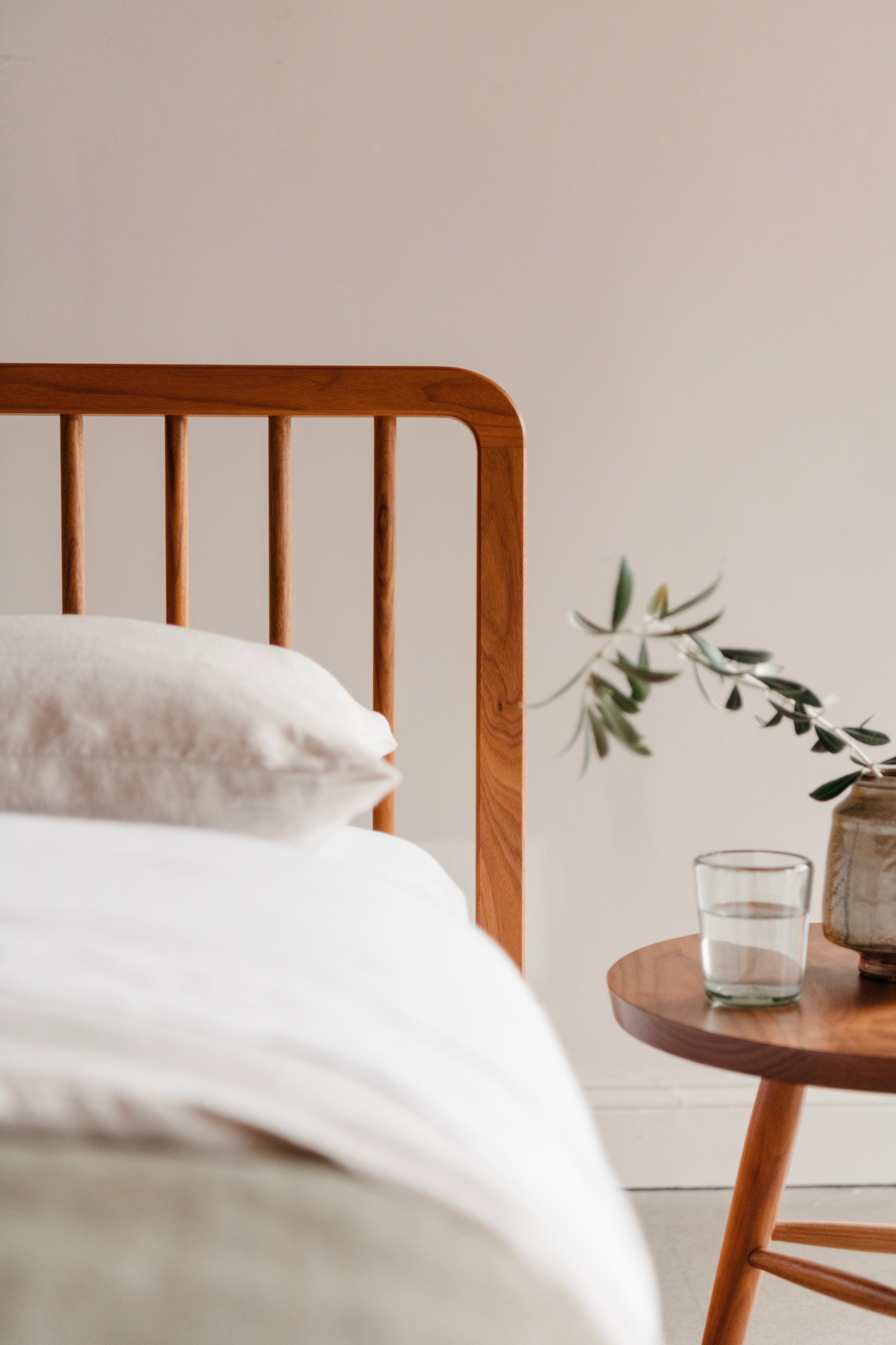 Close up of spindle headboard of walnut Chamberlain bed with white bedding and a small table with a glass of water and plant in the corner.