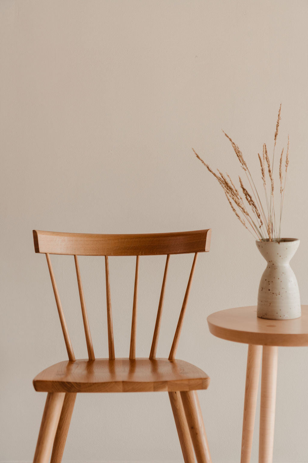 Wooden chair and small table with a vase of dried plants against a beige wall.