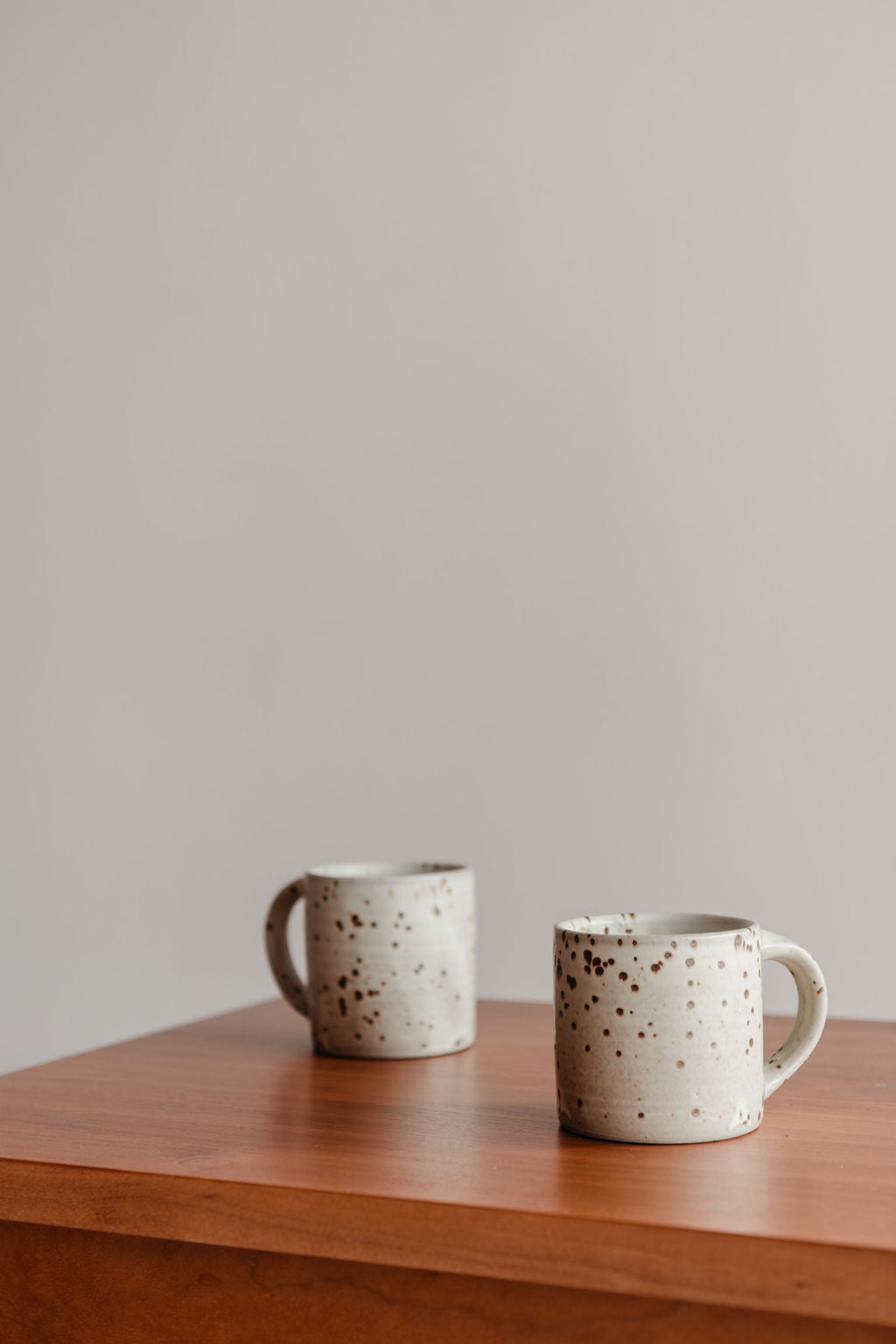 Two ceramic mugs with speckled pattern on a wooden surface against a plain wall.