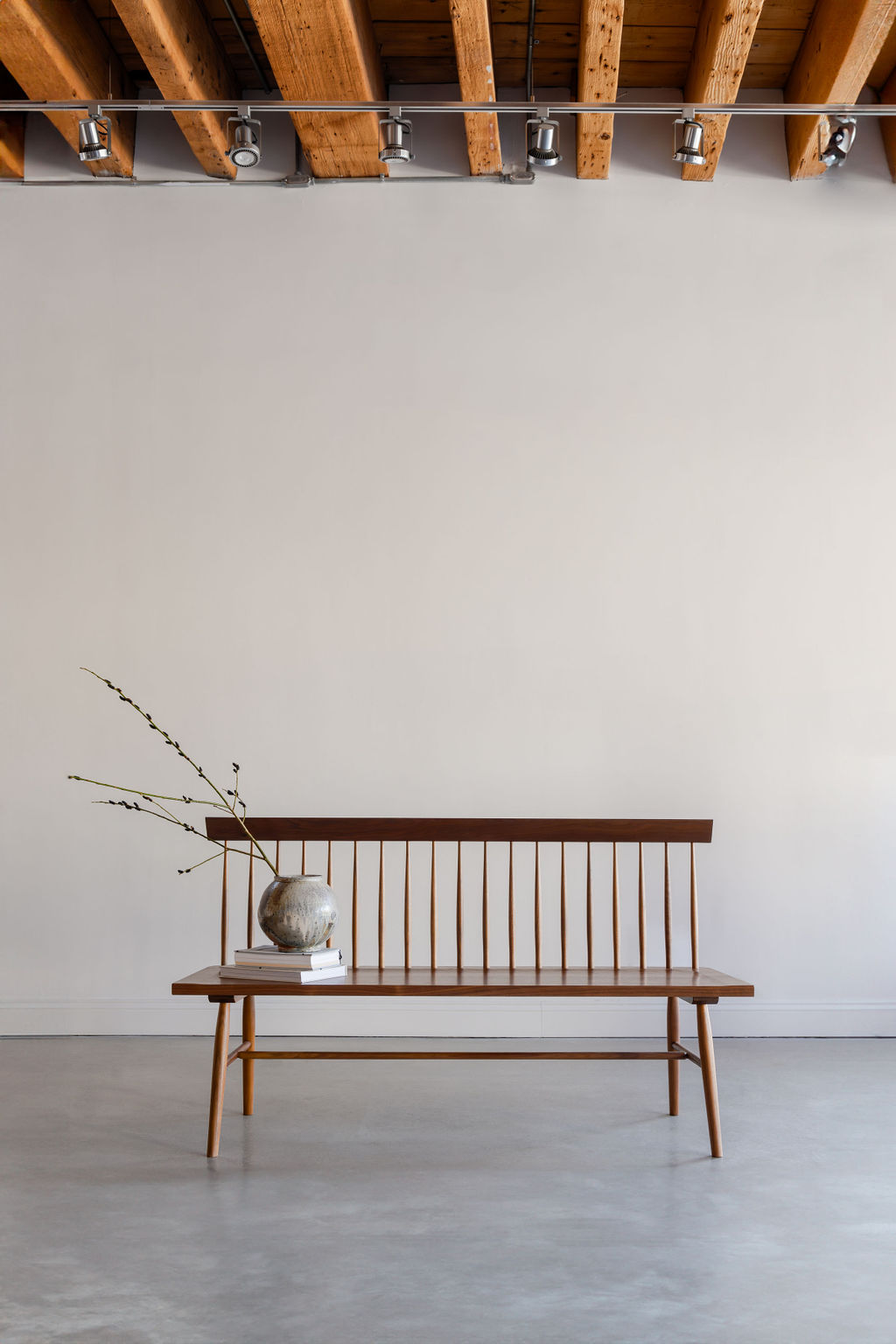 Head on view of the traditional Shaker style spindle back Broadside Bench in a modern room with wooden ceiling beams, cement floor and styled with pottery on books
