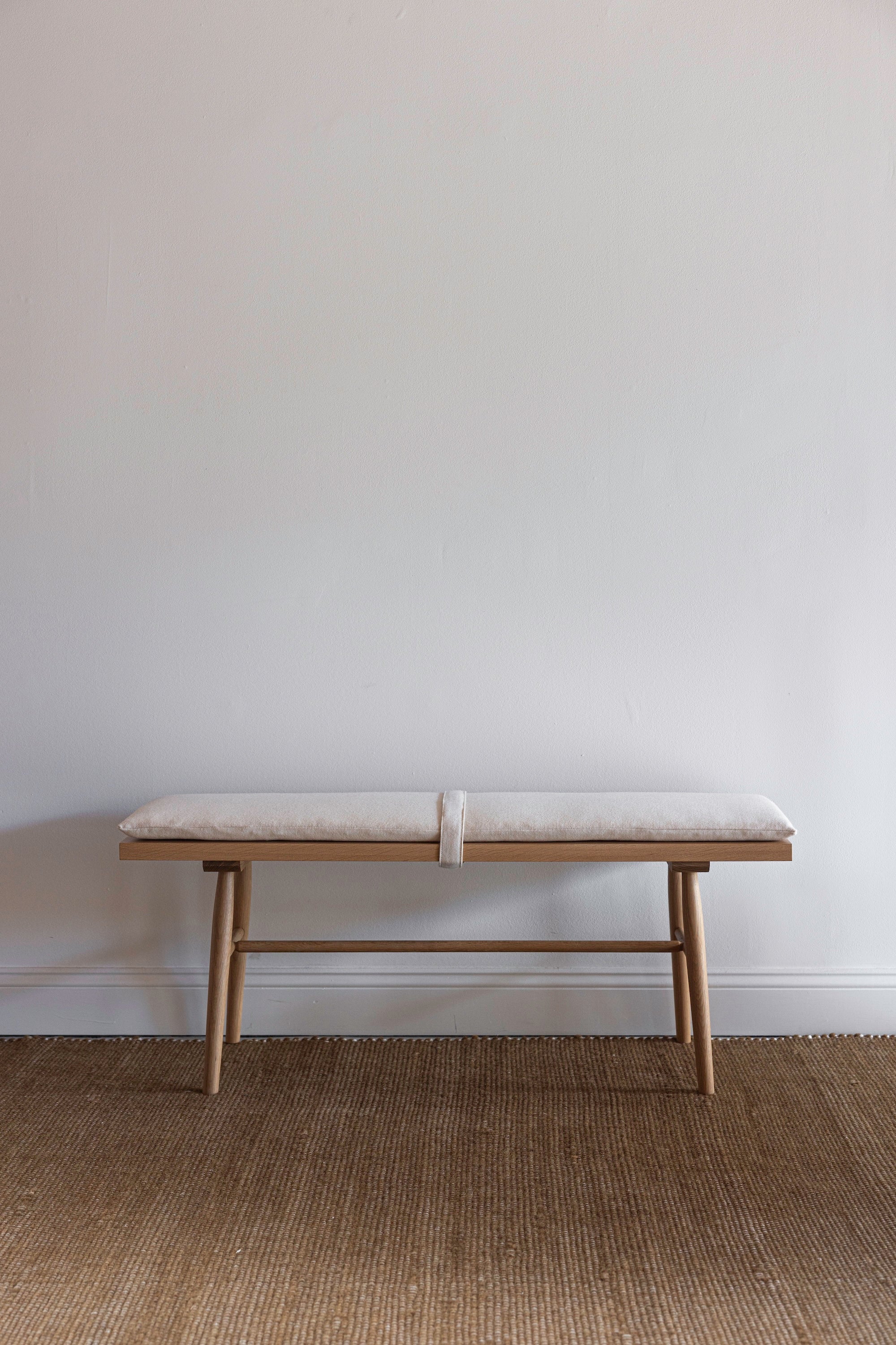 Wooden bench with round legs and a white cushion on top against a white wall on a brown rug
