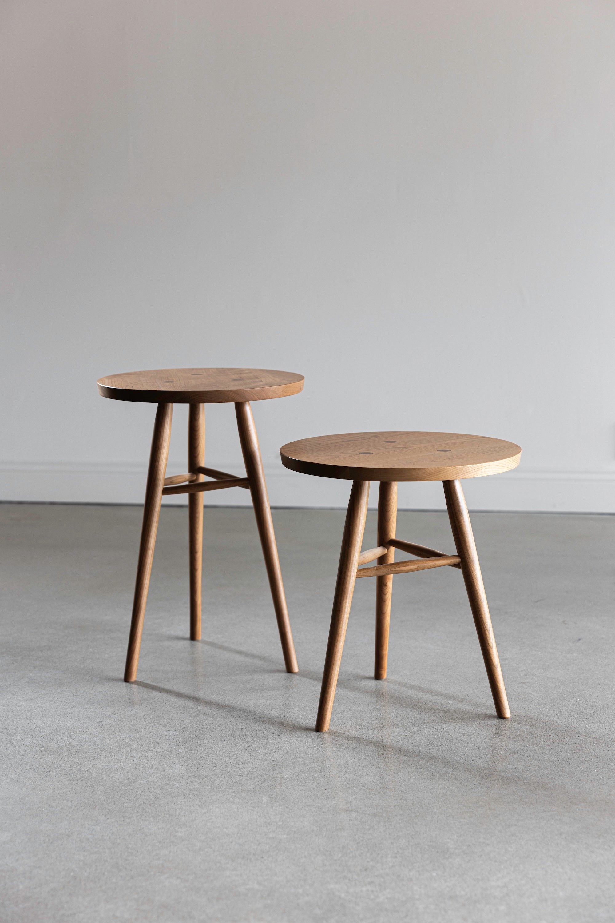 Two round wooden Hudson Side Tables with three legs in a plain room with cement floors and white walls.
