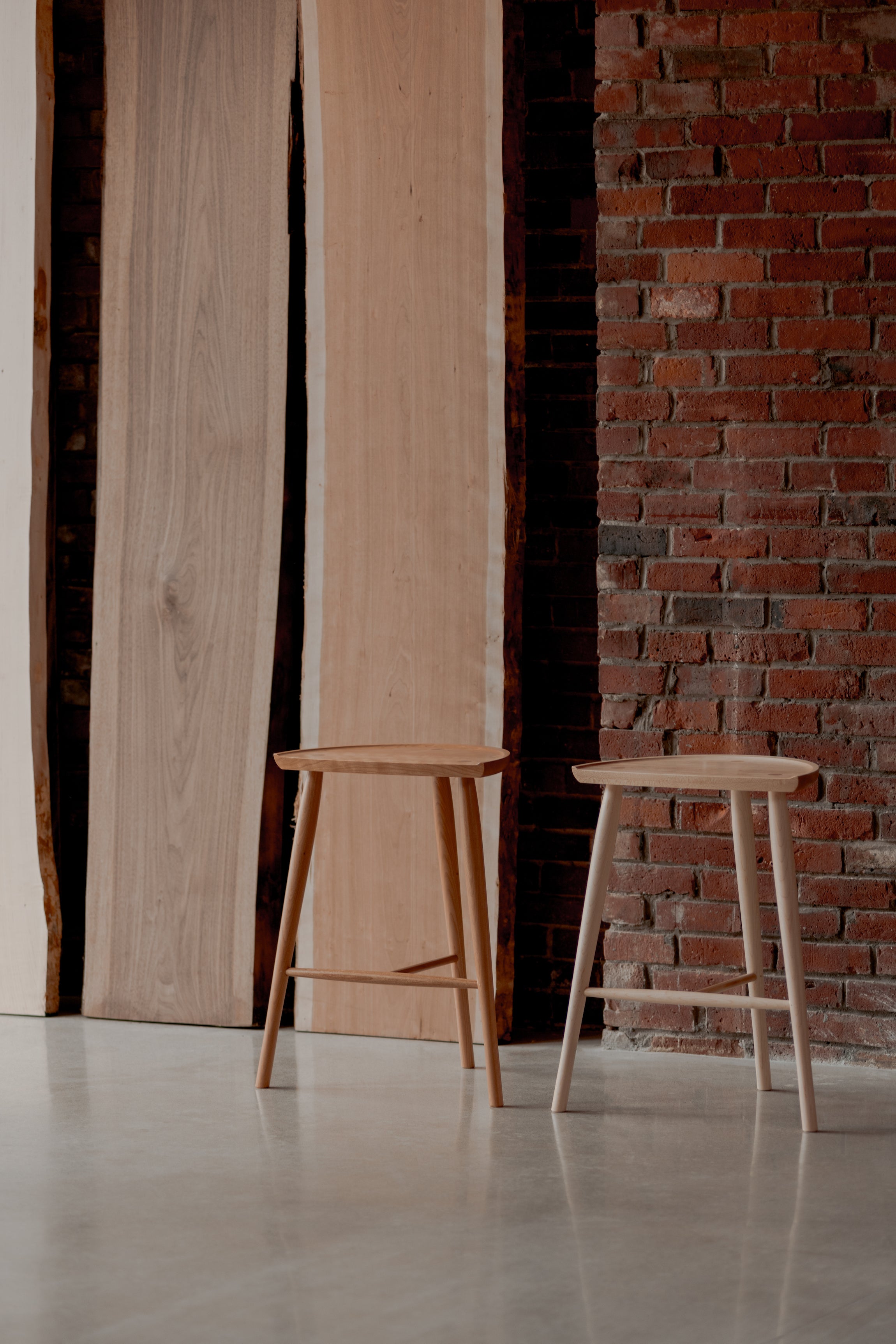 Two Shaker Stools with three legs next to brick wall and large slabs of solid hardwood