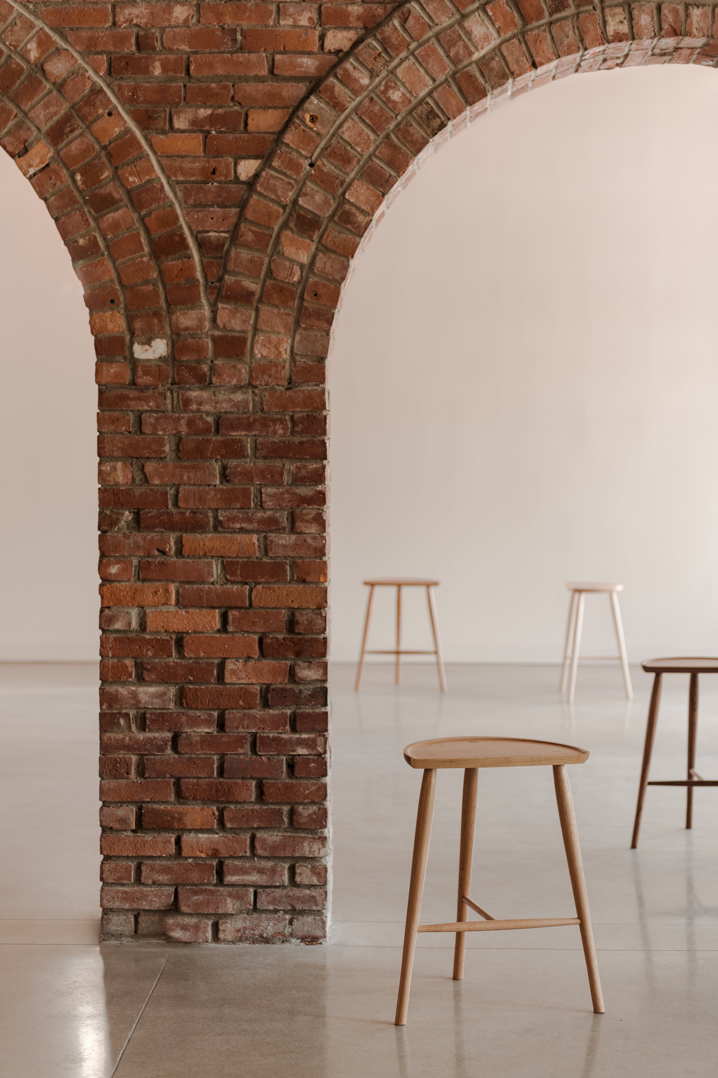 Four three legged Shaker stools built in different woods placed at varying distances on cement floor under brick arch architecture