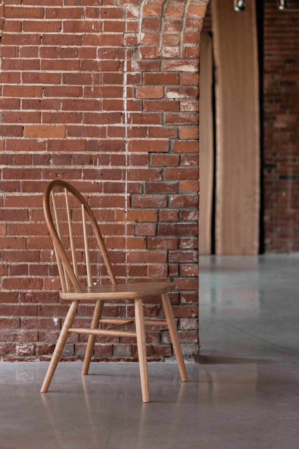 The bowback Hudson Dining Chair in front of a red brick wall with tall wood slabs in the background