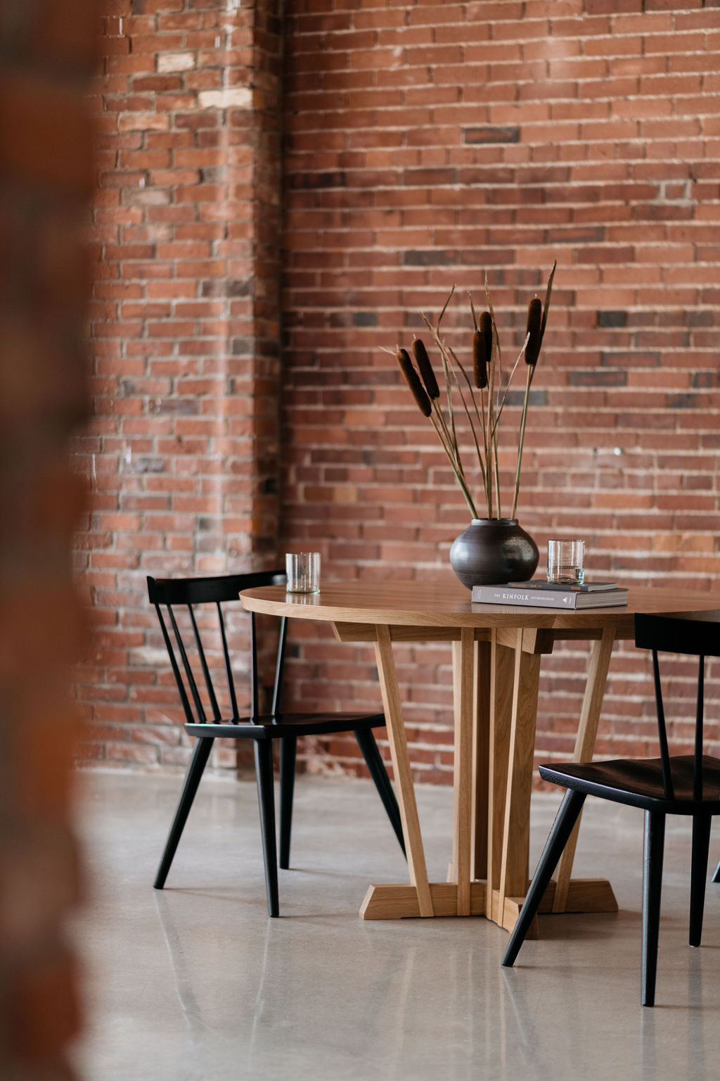 A round pedestal dining table in a large red brick room with two black Windsor chairs, two glasses of water, cattails in a vase and a book.