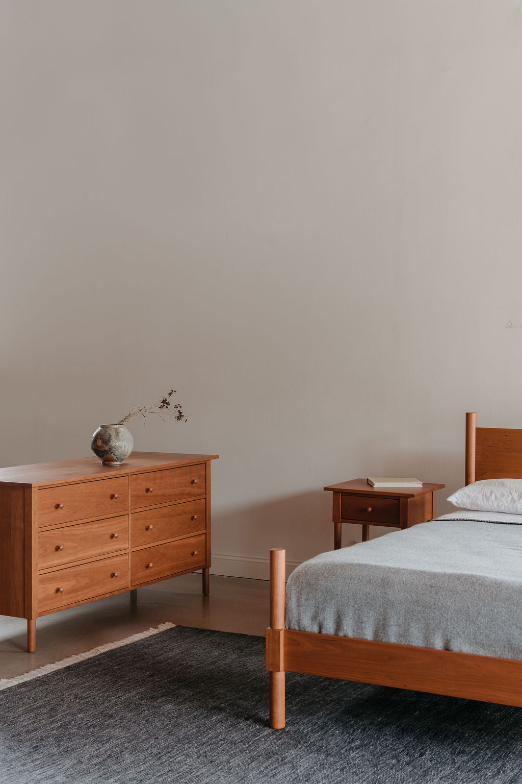 MS1 Wooden dresser and bed in a minimalistic bedroom with a plain wall.