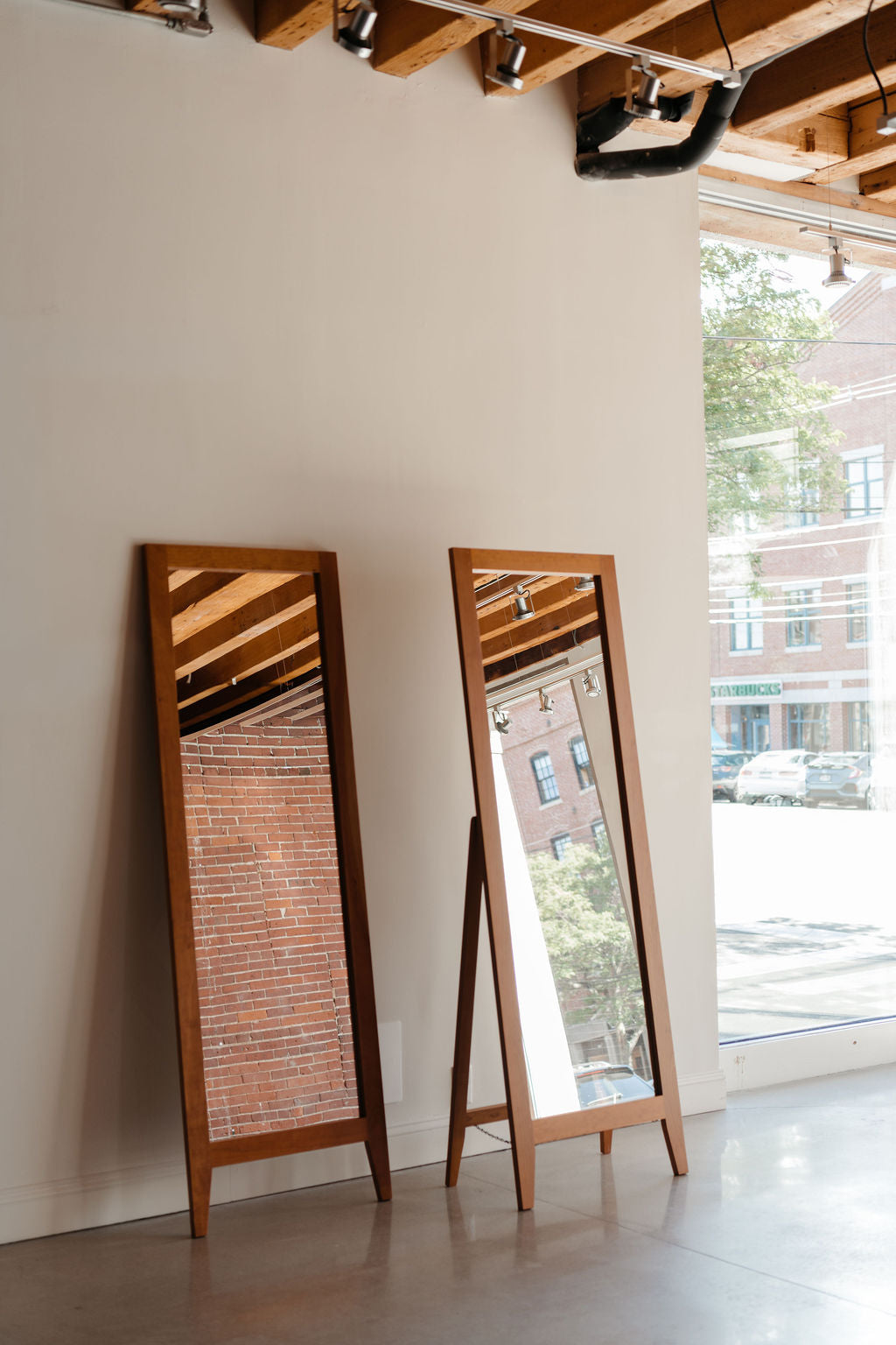 Two wooden floor mirrors against a white wall with a window in the background.