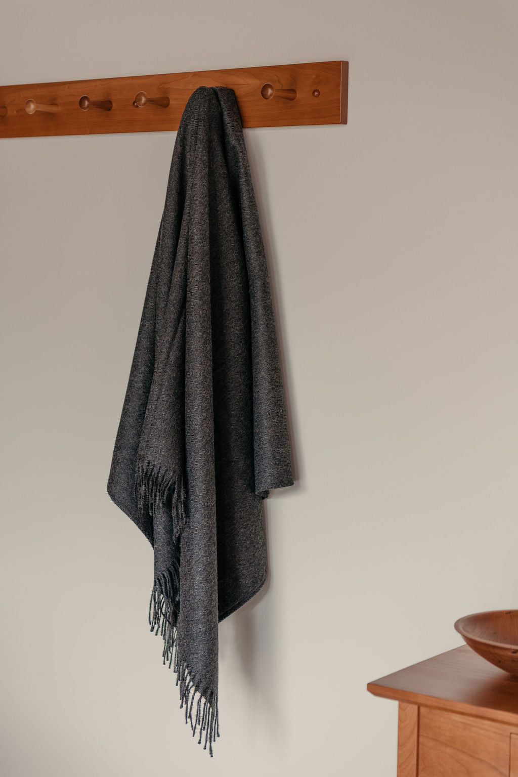 Gray blanket with fringes hanging on a wooden peg rack against a beige wall.