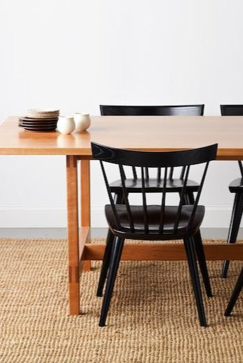 Ceramic plates and cups on modern cherry trestle table with four black modern windsor stye Boston chairs on beige woven rug with white background