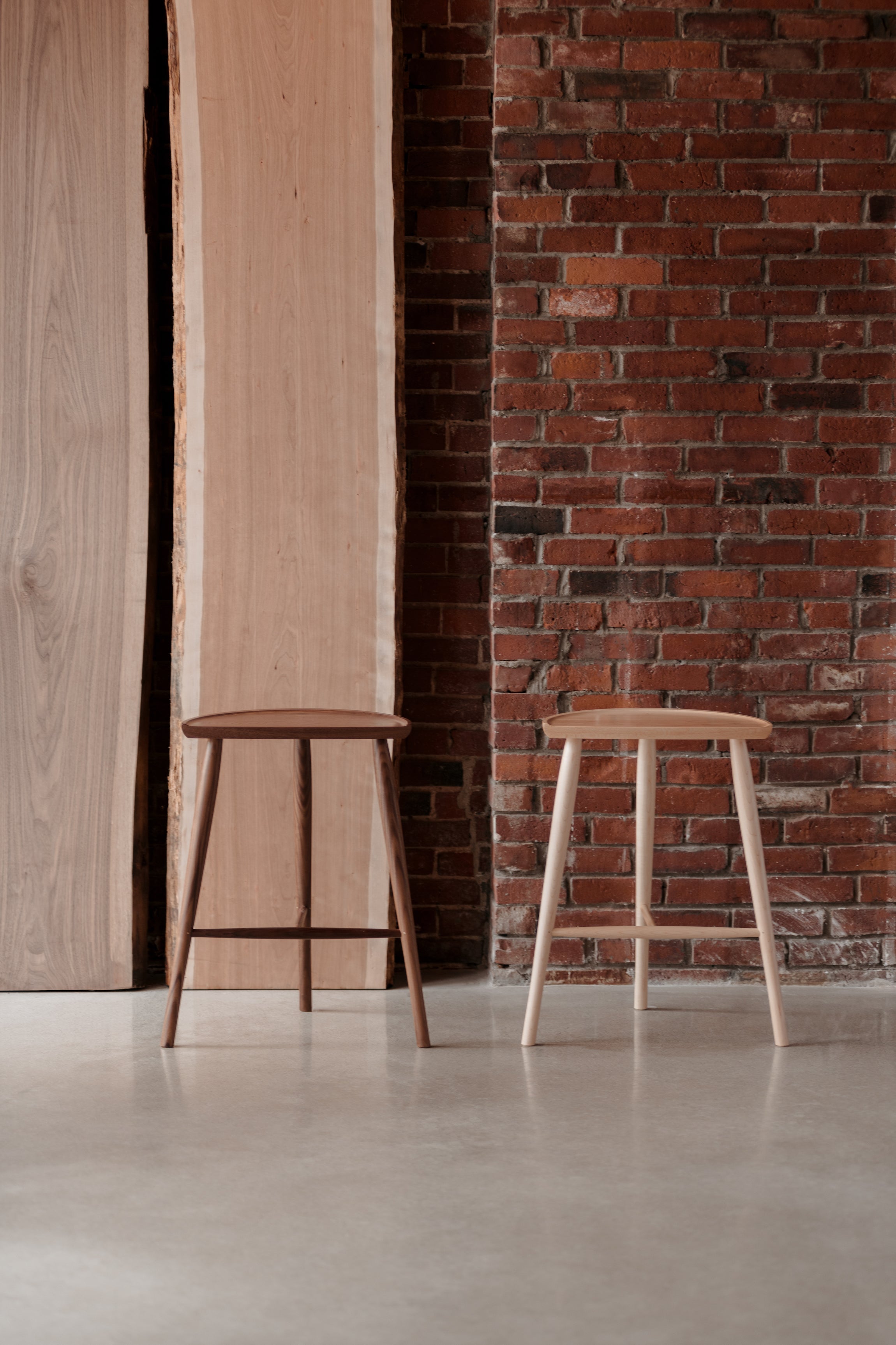 Two Shaker Stools with three legs next to brick wall and large slabs of solid hardwood