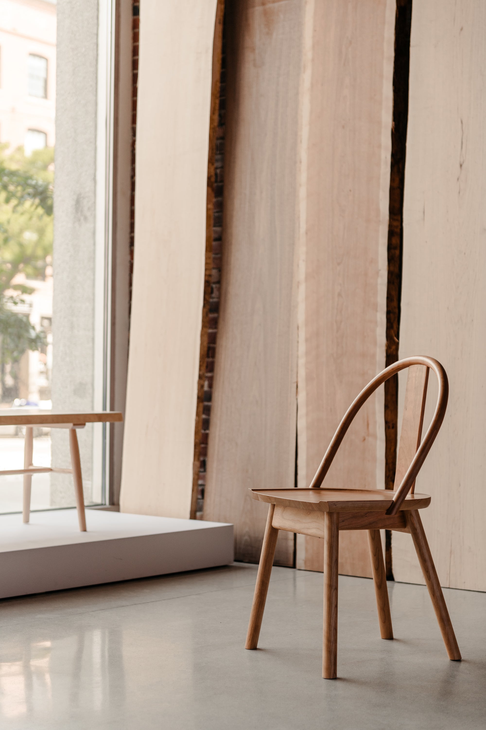 The Atlas Dining Chair from Arkansas's Mazhil & Co. in the large glass window of the Portland showroom next to large slabs of wood