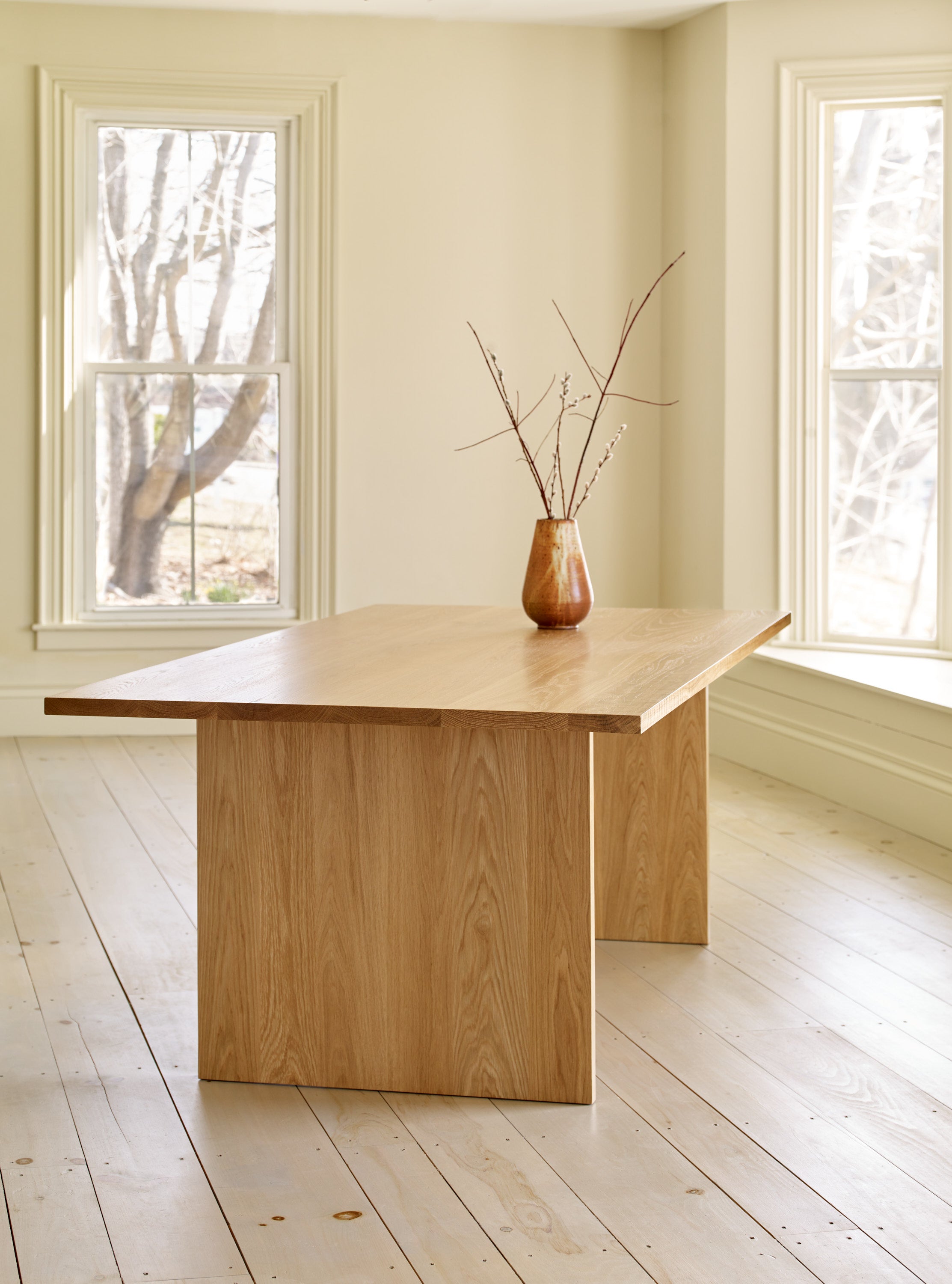 White oak trestle style Hygge Dining Table in bright window lit room on wood pine floors, styled with pottery vase