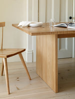 Minimalist dining photo with white oak table and windsor chair set with white plate, napkin, glass of water and a book in front of beige door on pine floor