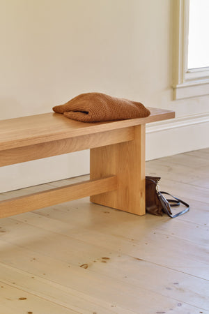 Modern trestle style Hygge Bench in white oak in bright room along wall with windows styled with rust orange sweater and brown purse on the floor