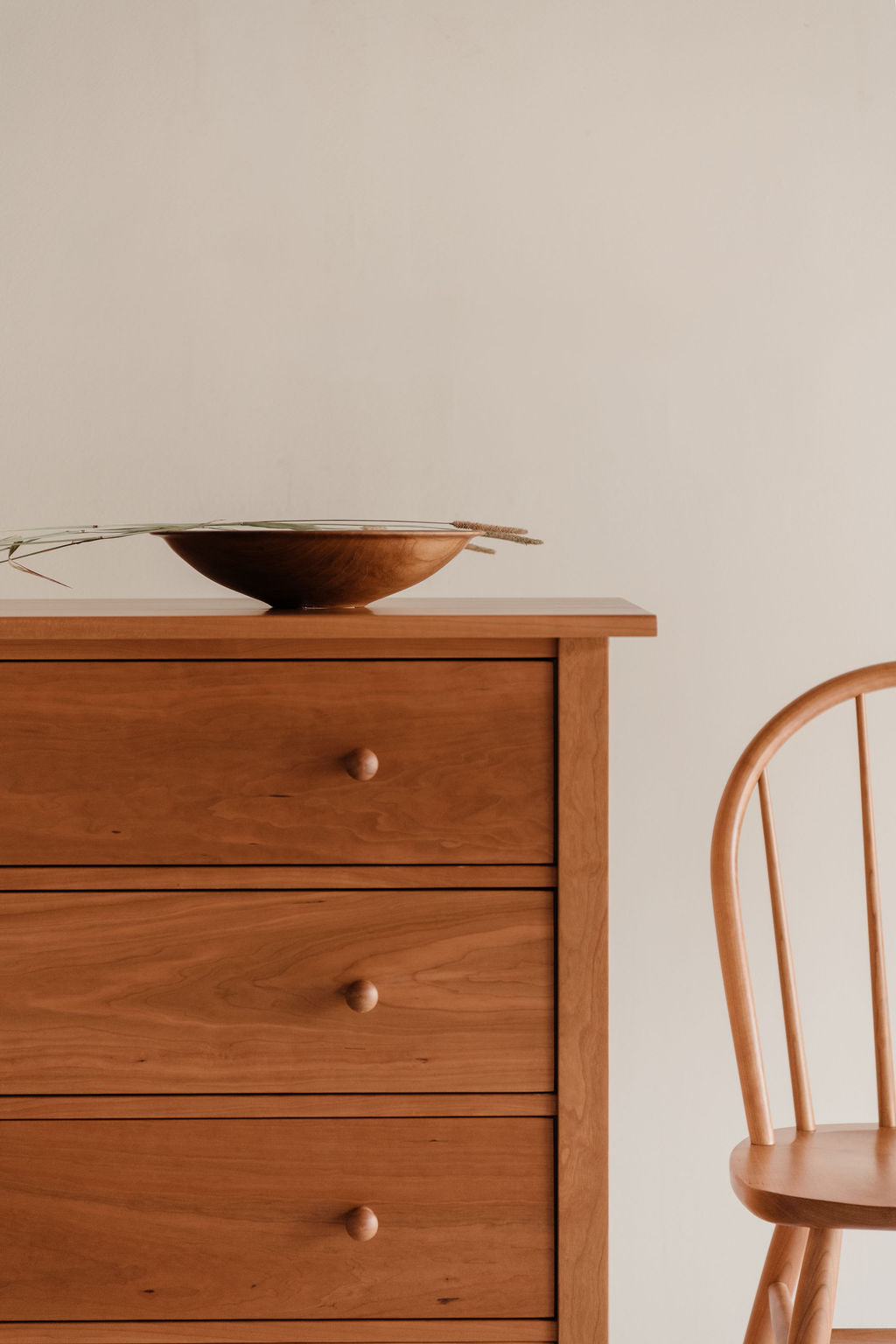 Wooden dresser with a bowl on top and a wooden chair to the side against a beige wall.