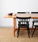 Ceramic plates and cups on modern cherry trestle table with four black modern windsor stye Boston chairs on beige woven rug with white background