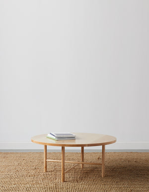 Round, maple wood, Scandinavian inspired coffee table with round legs on beige woven rug with table top magazines and white background