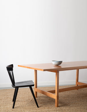 Ceramic bowl on modern cherry trestle table  with visible joinery with black modern Boston chair on beige woven rug with white background