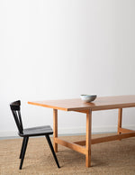 Ceramic bowl on modern cherry trestle table with black modern windsor style Boston chair on beige woven rug with white background