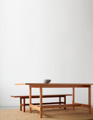 Modern trestle table and bench in solid cherry with pottery bowl on beige woven rug with white background