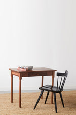 Small walnut MS1 desk with rounded legs, books on top, and one drawer paired with modern modern spindle chair