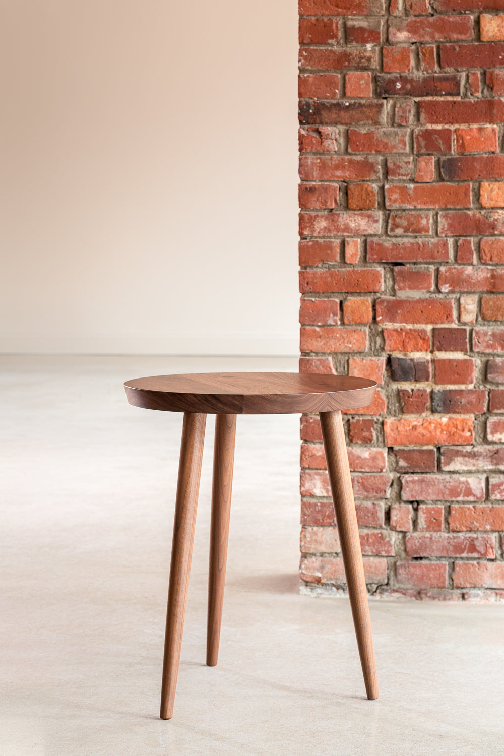 Modern round walnut nightstand with three round tapered legs along brick wall