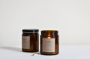 Two candles in amber glass jars, one with black lid and other with lit flame and burnt candle 